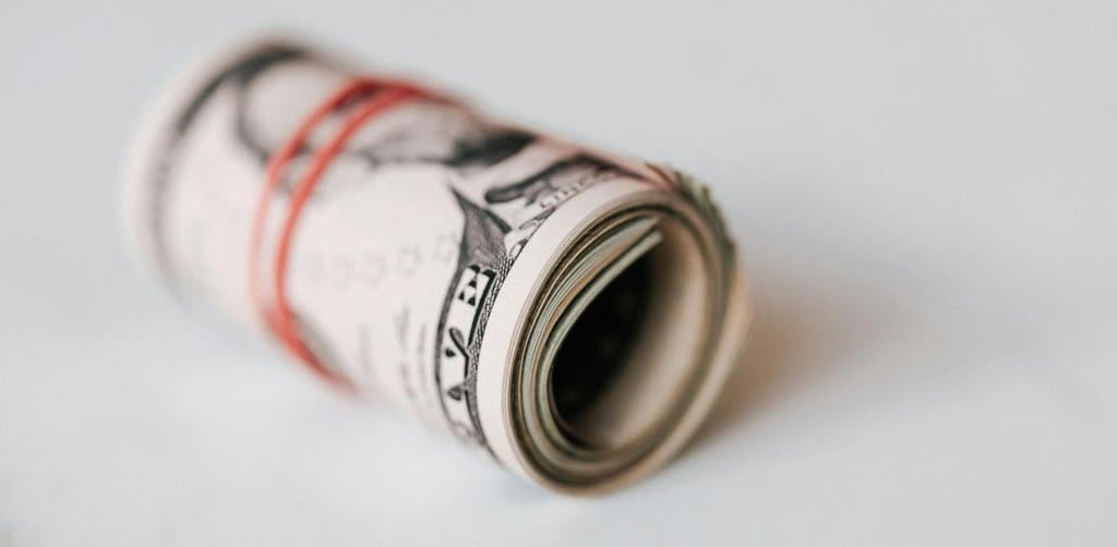 A bundled roll of US one-dollar bills tightly secured with a red rubber band lying on a clean white surface in soft natural light, the income yield that business note investing produces month after month for disciplined investors