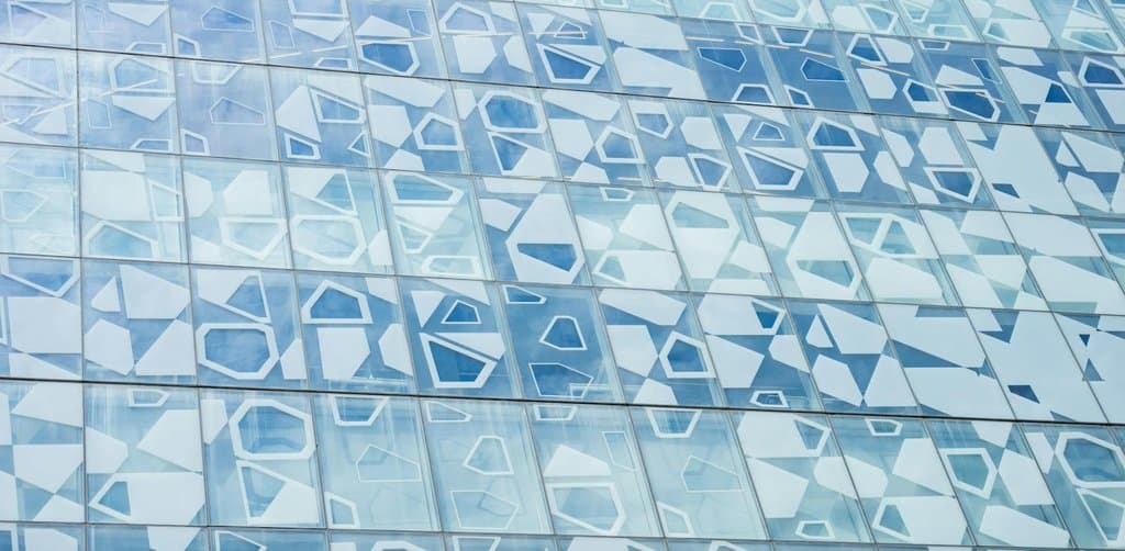An abstract architectural view of a tall building facade composed of dozens of glass panels arranged in pentagonal and triangular geometric patterns with white outlines against pale blue sky reflected in alternating cool and bright squares, the visual metaphor for the diversified portfolio structure that disciplined business note investing depends on