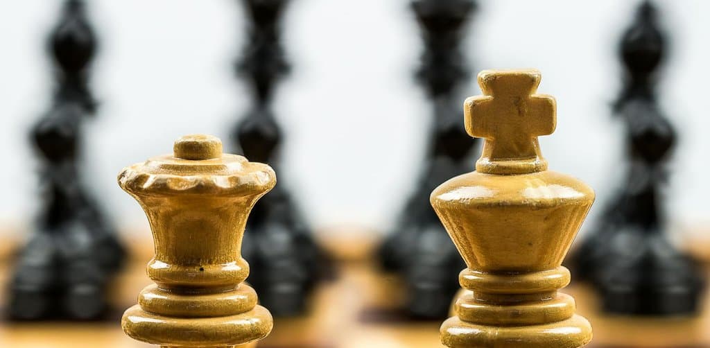 A close-up of two natural cream-colored wooden chess pieces in the foreground a king and a queen positioned together at the front edge of a polished wood chessboard with four dark black wooden chess pieces visible blurred against a soft white background in the rear of the board including a tall king and three flanking opposition pieces shown in shallow depth-of-field composition, the strategic-decision metaphor for the employee stock ownership plan vs stock options choice every employee weighs when comparing equity programs