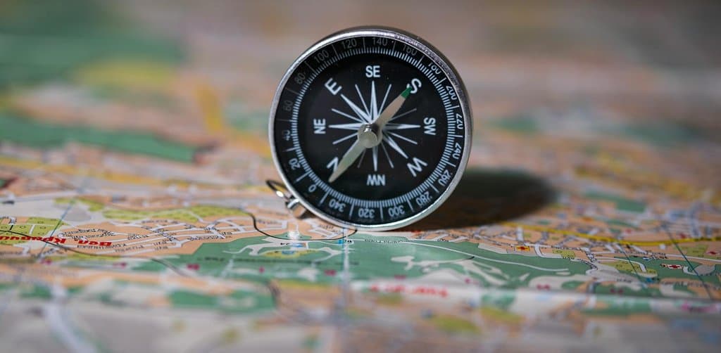 A close-up of a small round silver-rimmed pocket compass sitting on top of an open paper road map with the compass face showing white directional letters N E S W and degree marks around the rim with a green-tipped magnetic needle pointing toward southeast above a partially visible foldout green-and-orange topographic map with city street grids and roads in shallow depth-of-field, the orientation and decision-making metaphor for choosing between employee stock ownership plan vs stock options based on career stage and risk tolerance