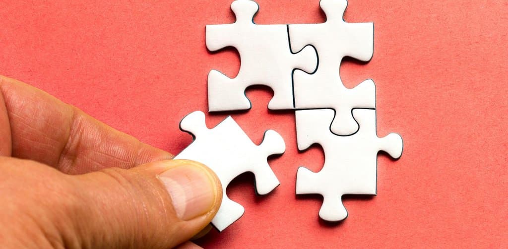 A close-up of three small white interlocking cardboard jigsaw puzzle pieces fitted together in an L-shape on a flat surface beside a fourth puzzle piece held between a thumb and fingertips approaching the cluster against a solid coral salmon pink colored paper background, the fitting-together metaphor for how an employee stock ownership plan vs stock options choice fits into a broader compensation and tax planning puzzle for every employee