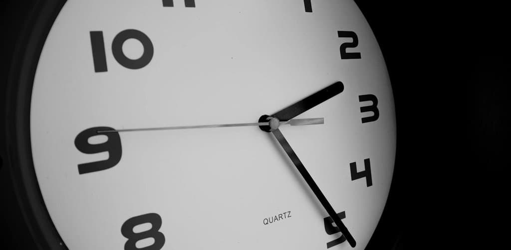 A close-up high-contrast monochrome image of a large round quartz wall clock with bold black numerals 1 through 12 arranged around the white clock face with the slim black hour and minute hands positioned at roughly twenty-two past two against a deep black background with the brand text QUARTZ visible below the center spindle, the time-bound waiting metaphor for the ERISA-mandated multi-year timeline an esop payout after termination follows before the participant receives the final payment