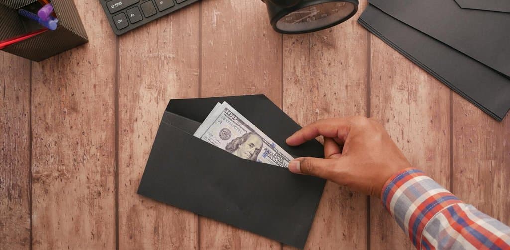 An overhead view of a wooden plank desk with rust orange and tan grain showing a hand reaching from the right edge to pull crisp green twenty-dollar U.S. dollar bills from inside a black paper envelope alongside the corner of a black computer keyboard with white labeled keys a black round alarm clock a second sealed black envelope at the right edge and a small black-and-yellow pencil holder with assorted pens at the upper left, the literal cash distribution payment metaphor for receiving the esop payout after termination via mail or direct deposit