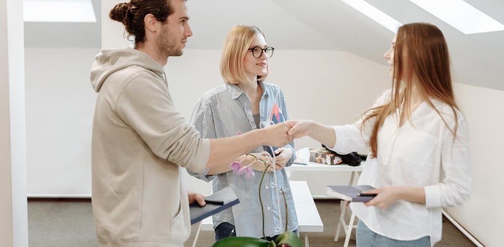 A natural sunlit office scene with three young professionals in casual business attire shown standing in a bright white-walled attic-style room with sloped angled ceilings featuring two angled skylights overhead with a small potted purple orchid on the central white table the man on the left in a beige hoodie holding a folio while the seated woman in striped shirt with glasses watches the standing woman on the right in a white blouse shake hands with him in a farewell departure handshake, the literal exit transition metaphor for the moment an esop payout after termination of employment is initiated by the plan administrator