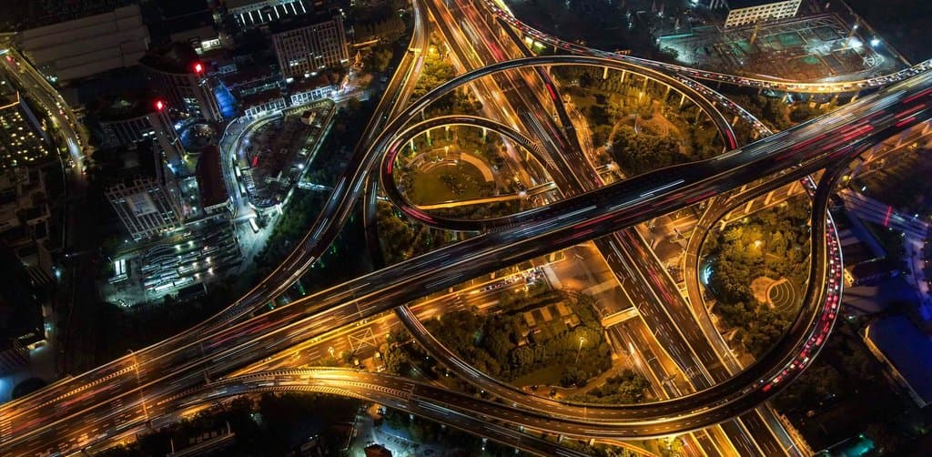 An aerial nighttime photograph of a multi-level highway interchange with curving overpass ramps illuminated by yellow streetlights and traffic light streaks weaving through dense urban surroundings of low-rise buildings and dark green park areas, the visual metaphor for the multi-channel pipeline a working note broker maintains to keep deal flow moving