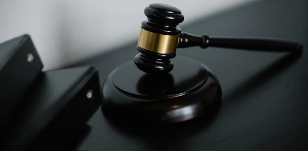 A polished dark wooden judge's gavel resting on its matching round wooden sound block with a brass band around the hammer head photographed on a black surface against a soft white wall background in moody side lighting, the symbol of the auction-style bidding process at the heart of how to broker business notes