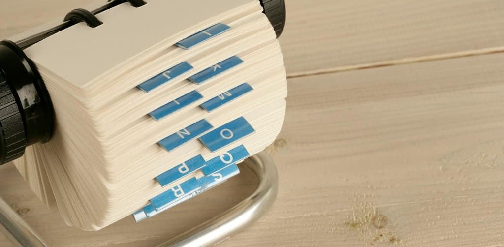A close-up of a vintage open Rolodex card file tipped on its side on a pale wood plank surface revealing a thick stack of cream paper cards with blue index tabs alphabetized from K through R protruding from the top edges, the institutional buyer Rolodex that every working note broker spends years building