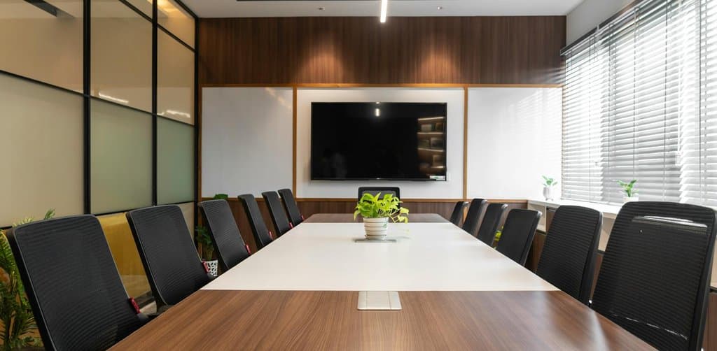 A modern empty boardroom with a long wooden conference table flanked by black mesh executive chairs, a wall-mounted television display, and floor-to-ceiling glass and wood walls letting in natural light through window blinds, the boardroom where leveraged buyout deals get structured and closed