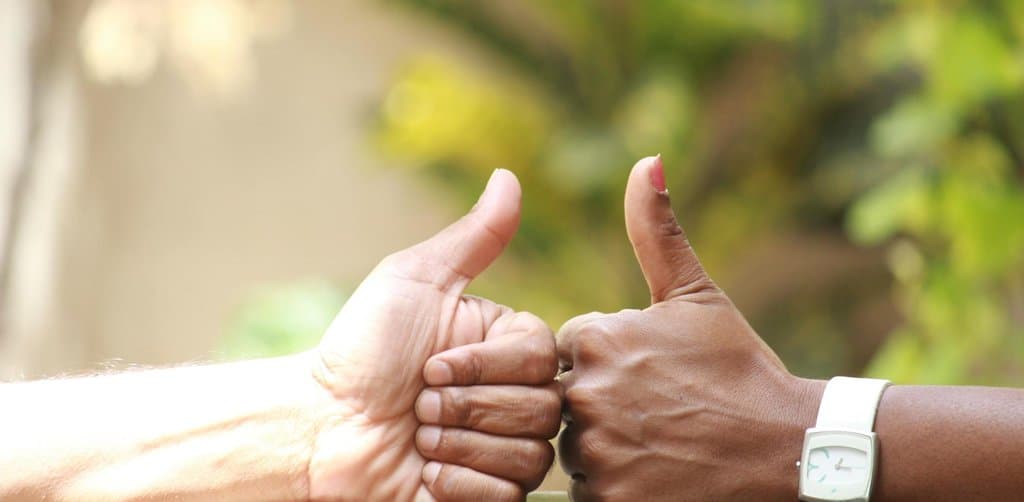 A close-up of two hands of different skin tones meeting in matching thumbs-up gestures resting on a wooden table edge against a blurred bright green leafy background, the simple positive symbol that masks the more complex pros and cons of seller carry financing