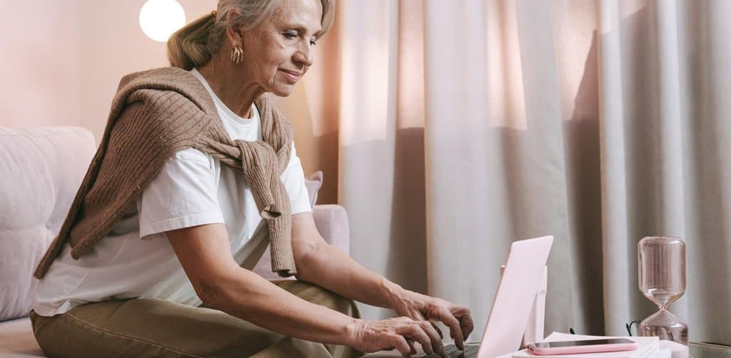 A silver-haired older woman in a white t-shirt with a brown cardigan draped over her shoulders sitting on a sofa working on a pink laptop with an hourglass and stacked books on the side table, the kind of seller weighing the pros and cons of seller carry financing during the retirement planning window