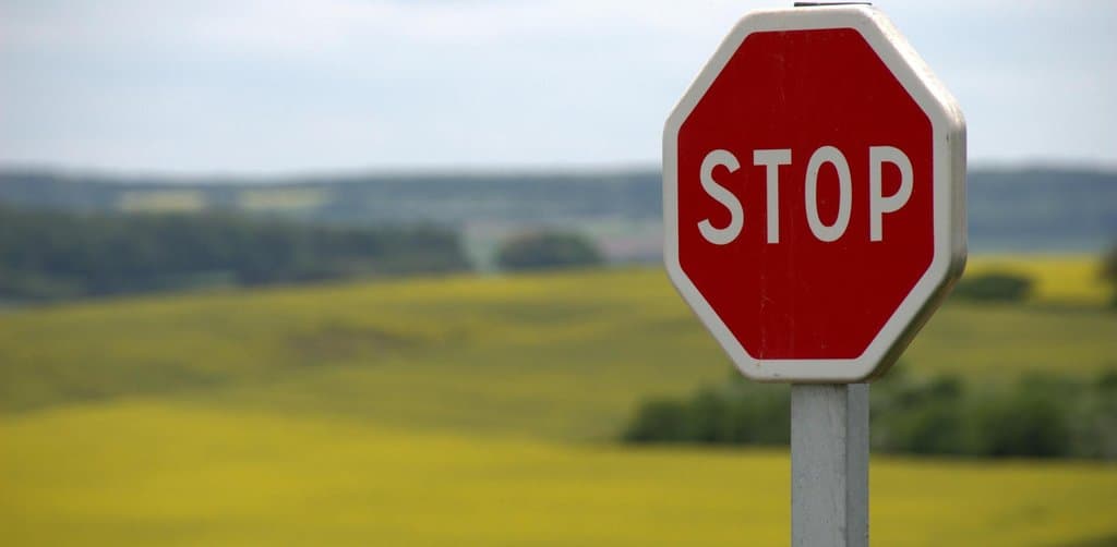 A red octagonal STOP sign mounted on a white metal post in the right foreground with a blurred yellow flowering rapeseed field and rolling hills under partly cloudy sky in the background, the cautionary signal that flags when the cons of seller carry financing outweigh the pros