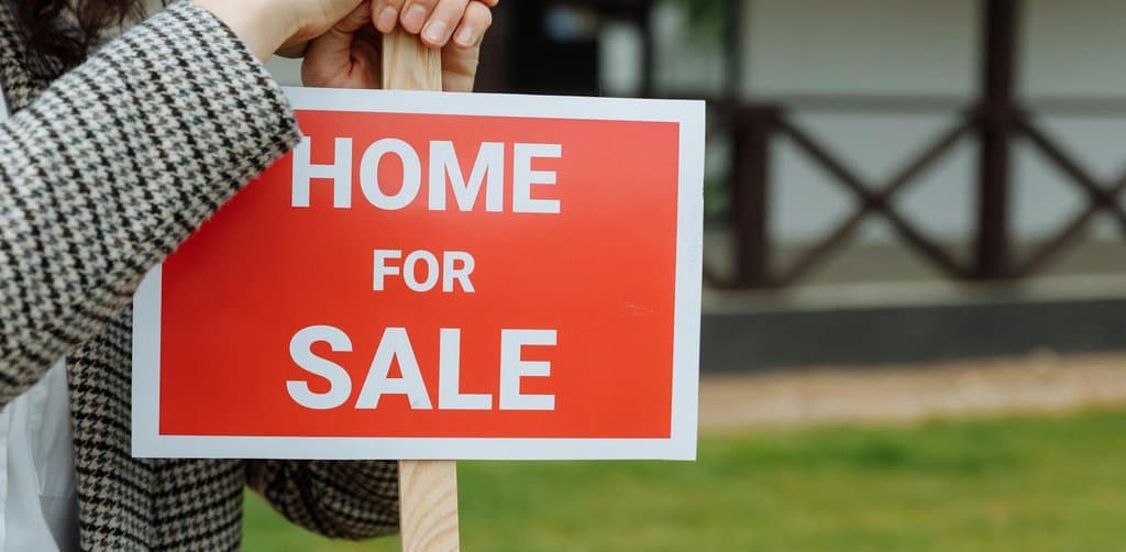 A close-up of a woman in a checked jacket holding a bright red rectangular HOME FOR SALE yard sign on a wooden stake against a green-grass lawn and blurred residential porch in the background, illustrating the moment an owner says sell my business and decides to take the leap