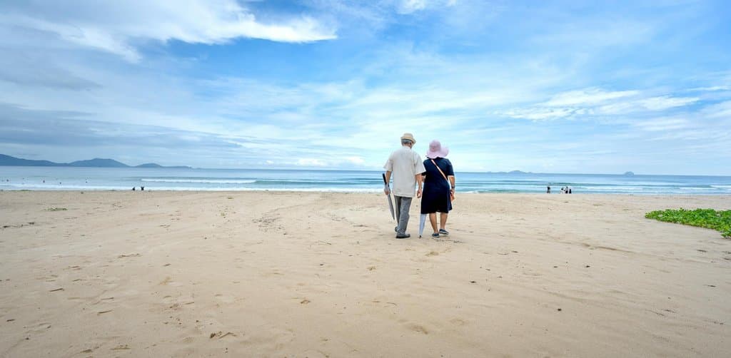 A bearded older man in light beige and a woman in a navy dress and pink sun hat walking along a wide empty white-sand beach toward the calm ocean horizon under a partly cloudy sky, the retirement life that the sell-my-business event makes possible