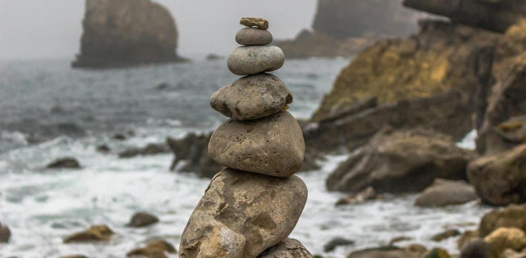 A carefully stacked cairn of rounded beach stones balanced from largest at the base to smallest at the top against a foggy ocean and rocky coastline backdrop, the balance metaphor at the center of every seller financing structure