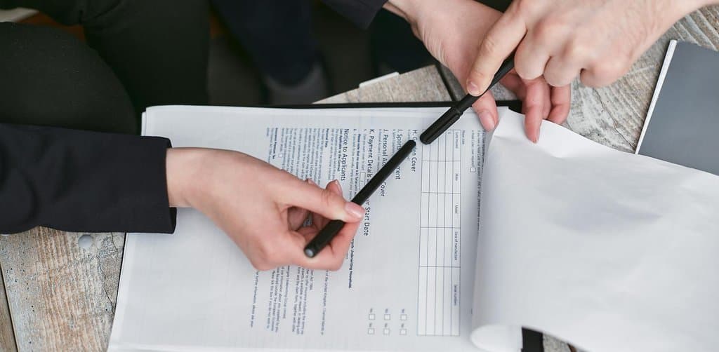 Top-down view of two business professionals' hands holding black pens and pointing at multi-page printed application paperwork open on a light wooden table, the careful section-by-section review every seller financing structure requires before signing