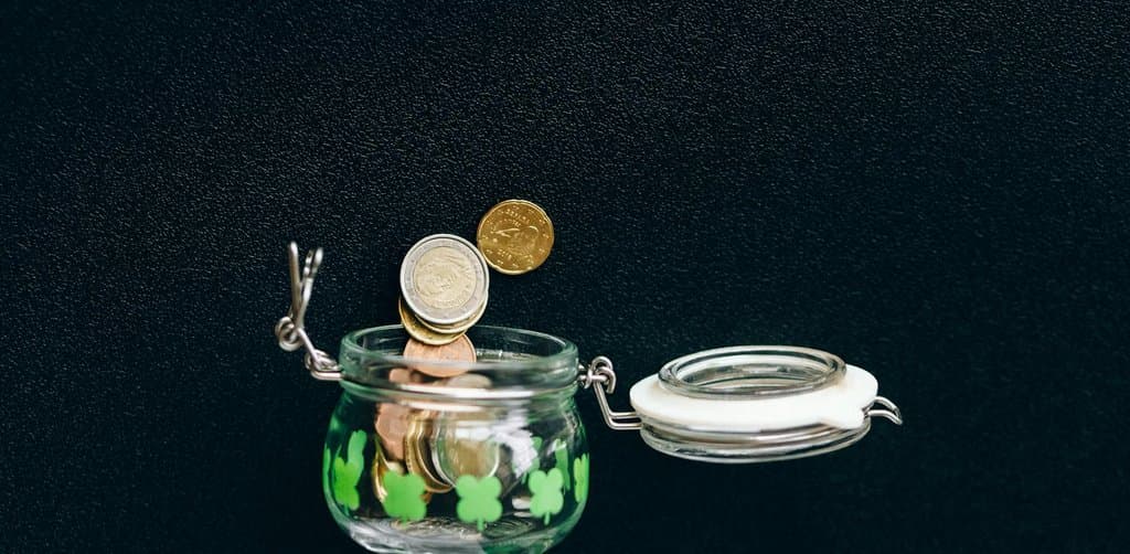 A small glass jar painted with green clover shapes filled partway with mixed coins and accepting a falling gold and silver coin from above against a black background, the gradual coin-by-coin growth that captures the contingent nature of an earnout structure