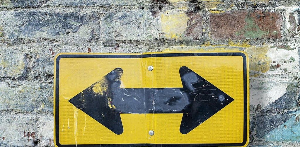 A bright yellow two-direction arrow road sign with one black arrow pointing left and one pointing right mounted on a metal post against a weathered painted brick wall with peeling paint, the directional choice at the heart of seller financing vs earnout