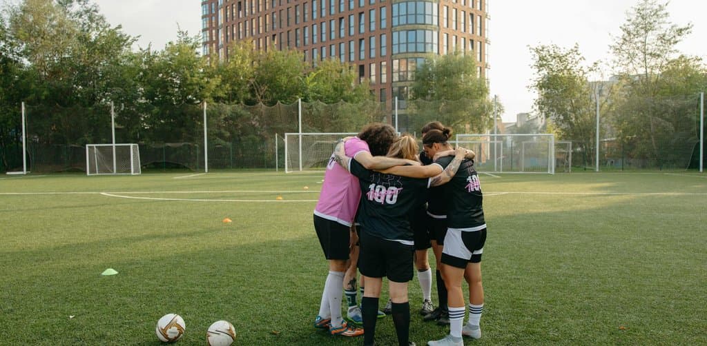 A women's soccer team in pink jerseys and black uniforms huddled together on a green artificial turf field with the player number 100 visible in front of city buildings in the background, the team-effort dynamic earnouts depend on for the seller to actually receive payment