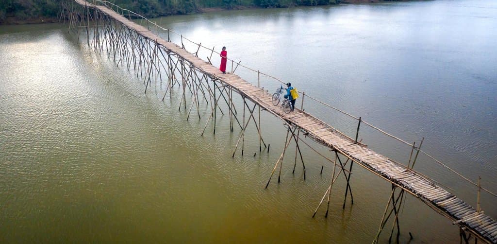 Aerial daytime view of a long narrow bamboo footbridge stretched on stilted wooden pilings across a wide brown river with one woman in a red dress walking toward the camera and a second figure with a bicycle near the middle, green farm fields visible on both riverbanks, a structural metaphor for the seller note bridging a transaction across a financing gap