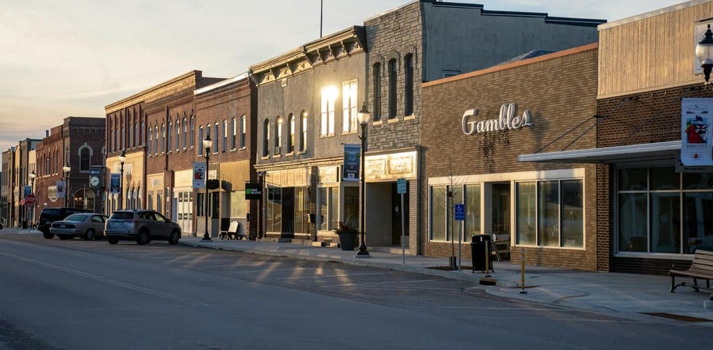 Quiet evening main street at dusk with brick storefronts on both sides of an empty two-lane road including a large brick building with cursive Gambles signage in golden window light, period-style street lamps casting long shadows on the pavement, the kind of small business district where seller-financed transactions keep ownership in local hands