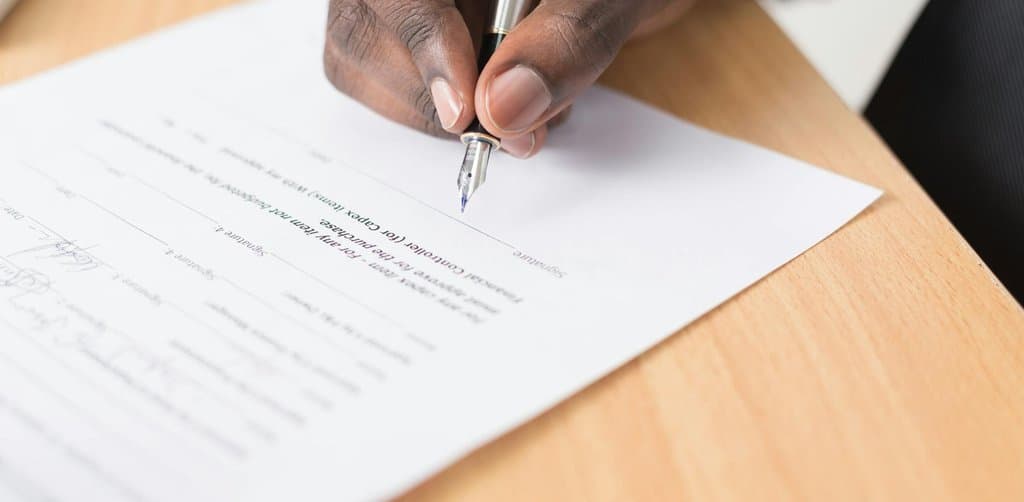 Close-up of dark-skinned hand in dressed shirt cuff signing a printed contract document with a slim chrome ballpoint pen on a light wood desk, the secondary signature line visible upside down at the top of the page, the moment a seller note becomes a real legal instrument