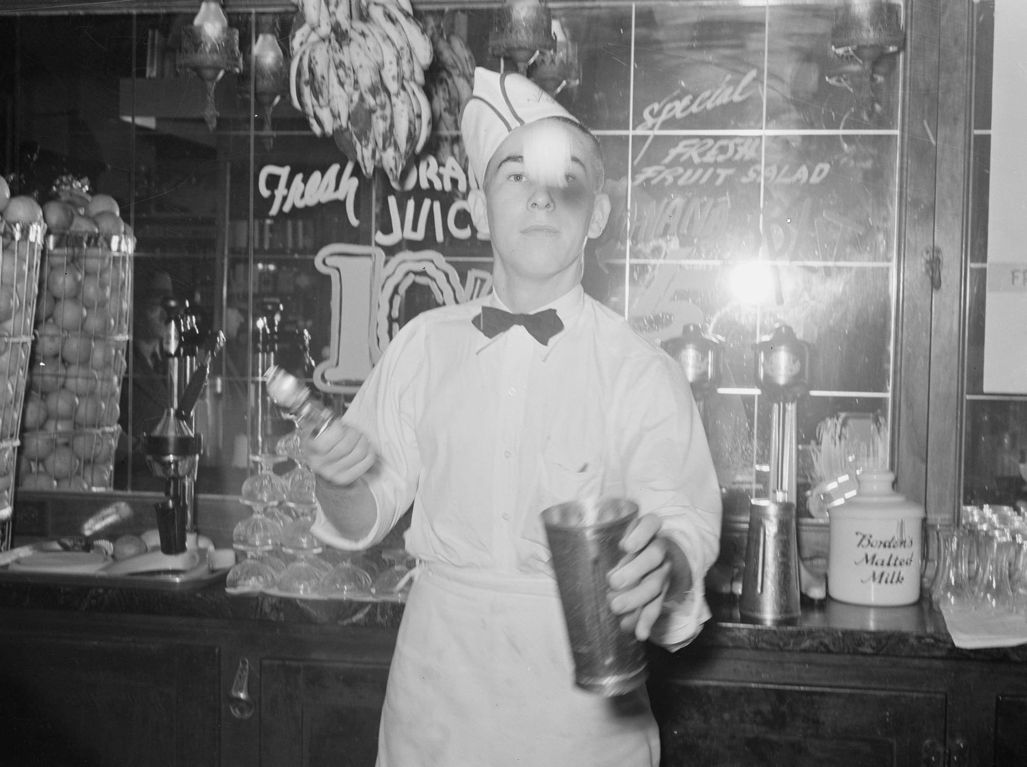 A soda jerk in a paper cap and bow tie tosses a shaker and a bottle over the counter of a soda fountain.