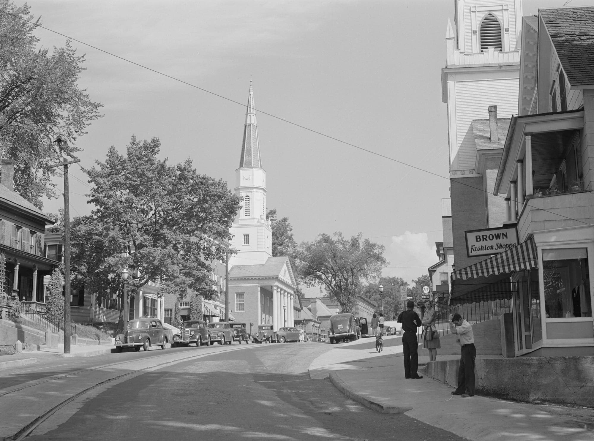 A hill street in a small Vermont town, parked cars along the kerb, a church spire ahead and a shop awning on the right.