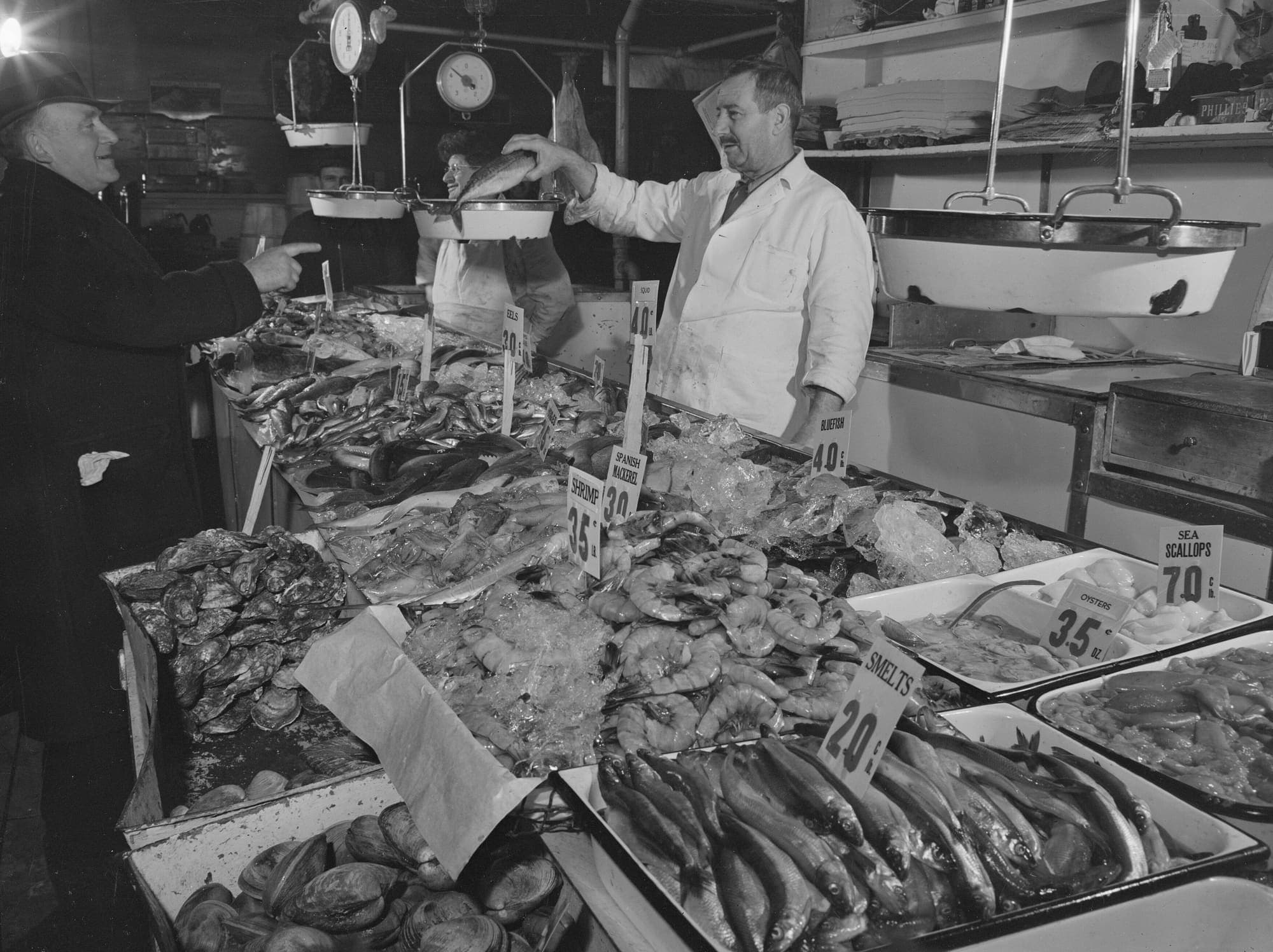 A fishmonger in a white coat holds a fish over his scales while a customer points at trays of shrimp, oysters and smelts.