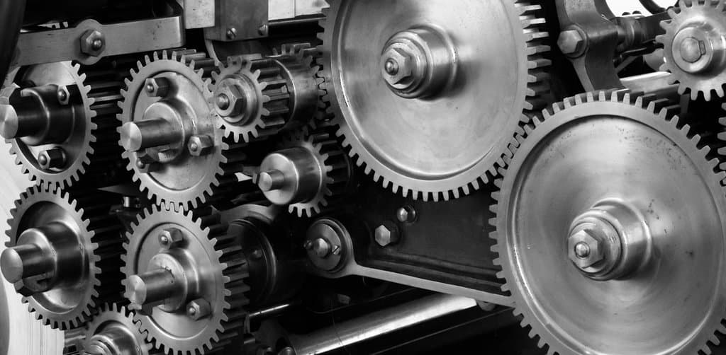 Detailed black-and-white photograph of a complex industrial gearbox showing multiple interlocking spur gears of varied sizes mounted on parallel shafts with visible bolts threads and grease marks across the steel surfaces, the mechanical interlocking that mirrors how an acquisition note interlocks senior debt and buyer equity inside a business purchase