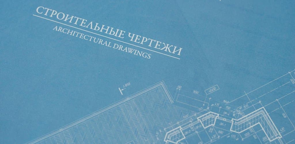 Close-up of a blue architectural drawings sheet on a desktop with bilingual title in white serif type and faint construction floor-plan linework with dimensions visible across the lower right corner, the planning blueprint metaphor for the structural acquisition note that completes a business purchase transaction