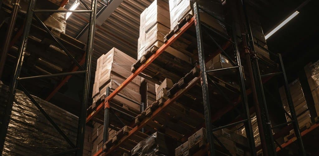 Vertical low-angle photograph of a dark industrial warehouse interior looking up at tall steel pallet racks loaded with shrink-wrapped pallets of cardboard boxes under overhead lights that catch the high ceiling beams, the visible business assets that secure the lien position behind an acquisition note in a business purchase transaction