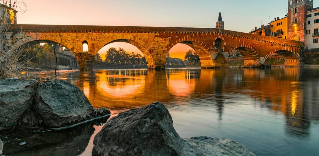 An ancient Roman stone arch bridge with four wide curved sandstone-and-brick arches reflected in calm reflective river water at golden sunset with smooth lit warm-toned medieval-Italian town buildings and a church bell tower visible on the far bank above the bridge and large weathered stones in the foreground rising from the river edge, the long-lasting durability and built-to-last metaphor for the multi-decade advantages of employee stock ownership plans that endure across owner transitions and generational shifts