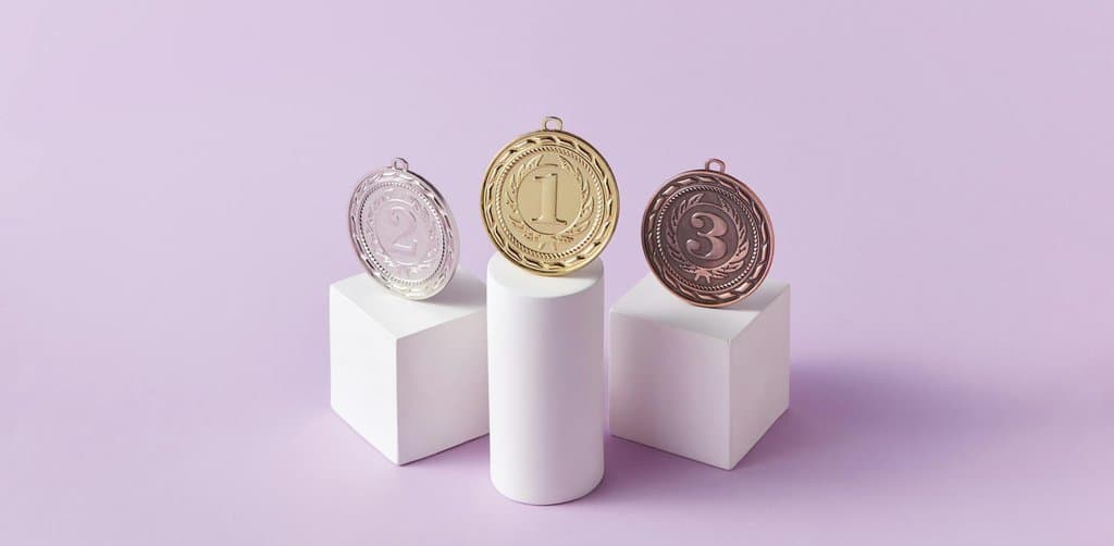 Three round metallic award medals labeled with numbers 1 in gold 2 in silver and 3 in bronze each propped up on individual white pedestal blocks of different heights with the gold first-place medal on the tallest center pedestal against a soft pastel violet-purple background in clean studio lighting, the structured competition-podium metaphor for the layered advantages of employee stock ownership plans that flow to owners workers and the company simultaneously