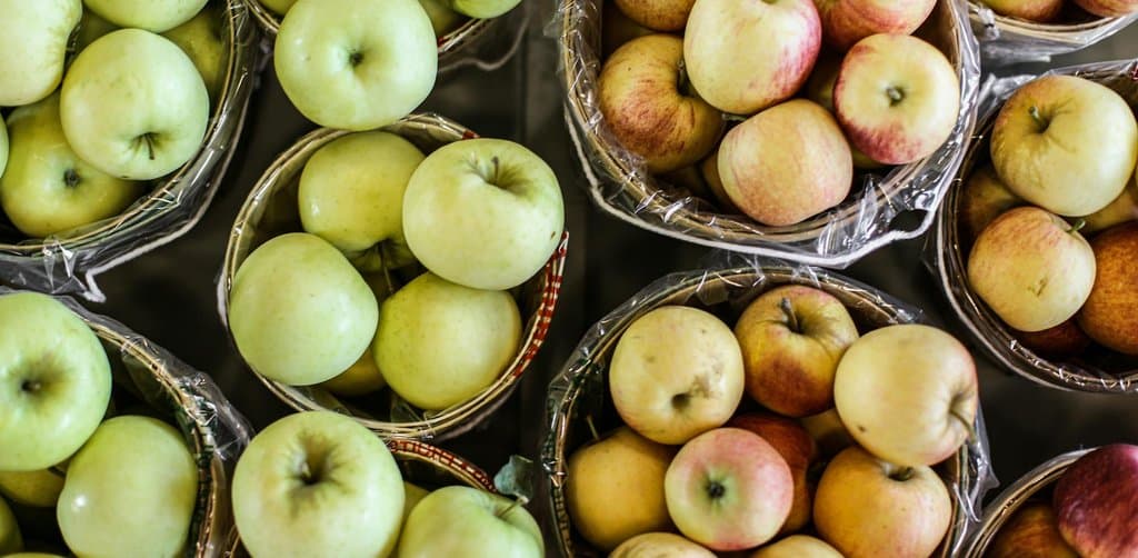 An overhead view of dozens of small light green and red apples filling four cellophane-wrapped circular bushel baskets arranged on a flat surface with the red apples on the right and the green apples on the left both varieties freshly picked and ready for market, the harvest abundance metaphor for the multiple stacked benefits of employee stock ownership plan participation an employee receives across a working career