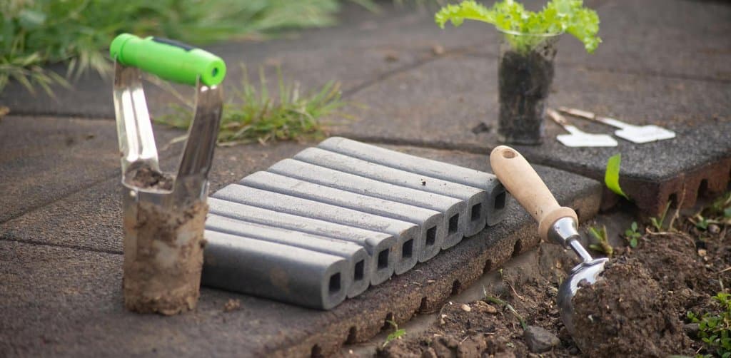 A stainless-steel garden trowel with a chrome bladed scoop and a green plastic handle stands point-up beside a stack of eight slim grey concrete bricks laid horizontally across a brick walkway with a brown wood-handled hand cultivator on the right edge and a small fresh lettuce seedling in a clear plastic seed cup with rooted dirt placed on the brick walk all in the soft afternoon shade of a backyard garden, the careful long-term cultivation metaphor for the cumulative benefits of employee stock ownership plan tenure across a working career