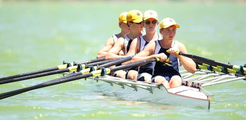 A men's four-person sweep rowing crew in dark navy unitards and matching yellow caps with crossed-blade insignia rows in synchronized stroke on smooth bright green lake water with all four pairs of yellow-tipped oars planted in identical position alongside the long slender white-and-black racing shell, the literal team alignment metaphor for how the benefits of employee stock ownership plan participation pull every participant in the same direction for the company