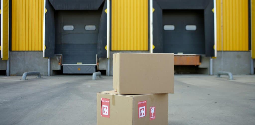 Two cardboard boxes stacked on the loading-dock pavement of a yellow industrial warehouse with two empty truck bays in the background, illustrating the inventory and infrastructure scaling that drives many real business funding reasons