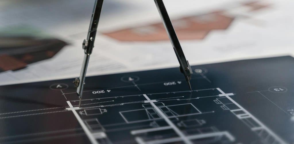 A drafting compass standing on a dark architectural floor plan with several blueprints in the background, the kind of planning artifact that defines the specific business funding reasons behind a serious capital raise
