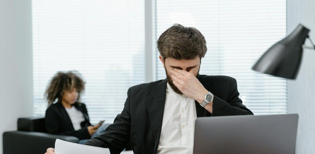 A bearded operator in a black blazer pinching the bridge of his nose while reviewing a financial document at a desk with a laptop and a colleague in the background, the cash-crunch moment that drives many of the most common business funding reasons