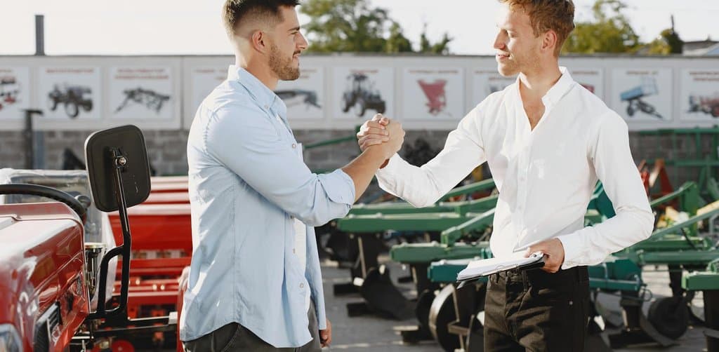 Two professionals shaking hands in front of farm and industrial equipment, the in-person handshake that often seals a private business investment