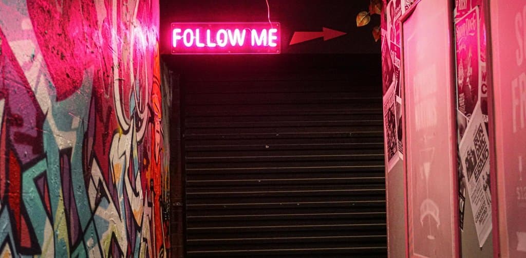 A bright pink neon FOLLOW ME sign with a magenta arrow points right above a dark staircase with vibrant graffiti street-art covering the left wall and red-tinted concert posters mounted on the right wall under a single overhead light bar at night, the literal directional sign metaphor for the clear decision point an owner reaches when choosing business loan vs personal loan