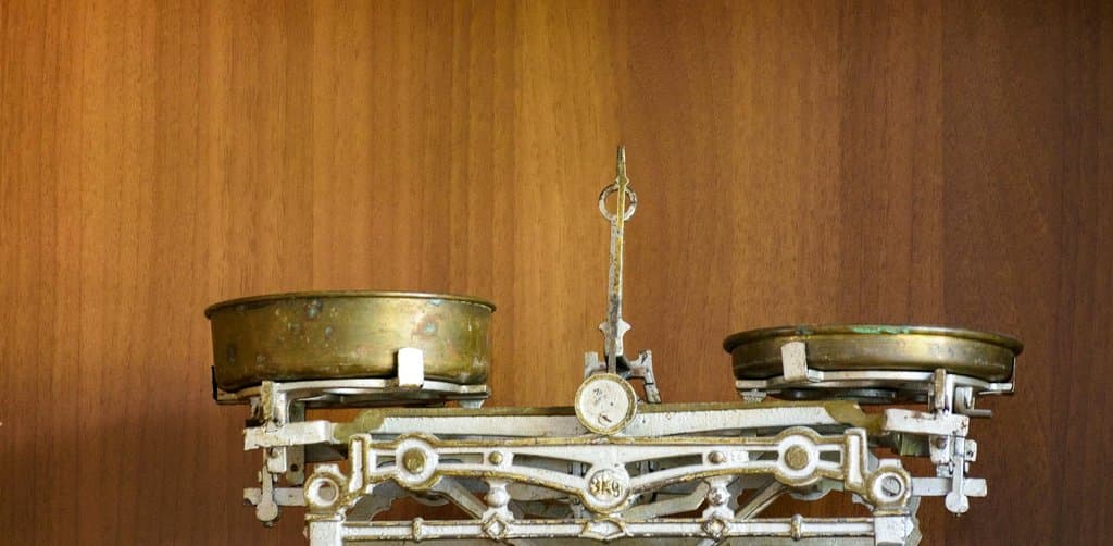 An antique brass-and-cast-iron two-pan balance scale with two empty round shallow brass pans sits on a wooden veneer shelf against a smooth medium-brown wood-paneled wall in even daylight with the pointer needle resting near center between the two arms, the literal weighing tool metaphor for the trade-off analysis behind a business loan vs personal loan decision