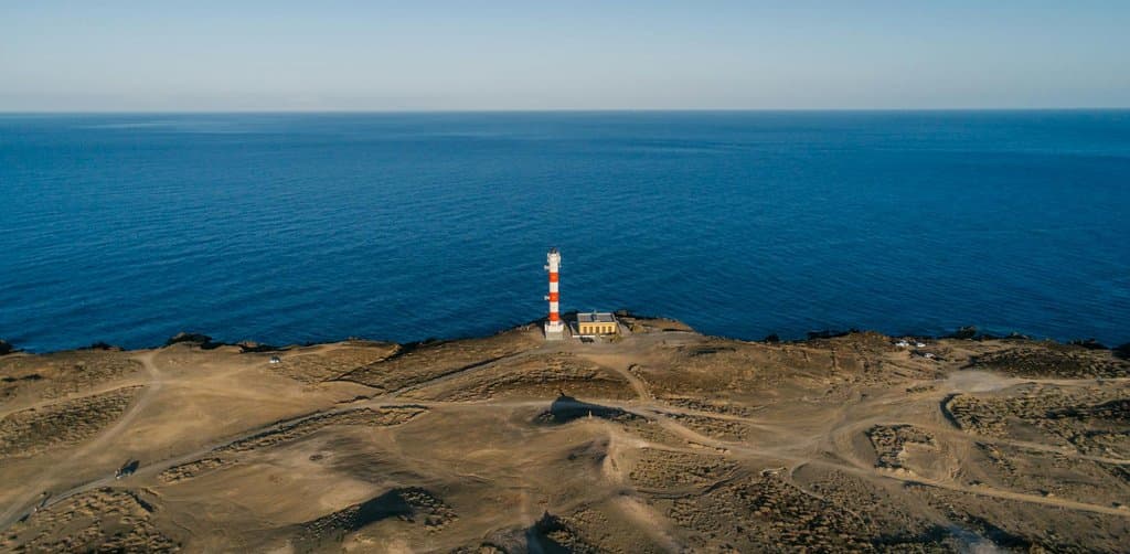 A small red-and-white striped lighthouse with a small attached caretaker's building sits alone on a wide pale desert headland of low scrub at the edge of dark navy ocean stretching to the horizon under a clear bright blue sky, an aerial daytime image of the singular guidance point a business note broker provides for sellers crossing the secondary market