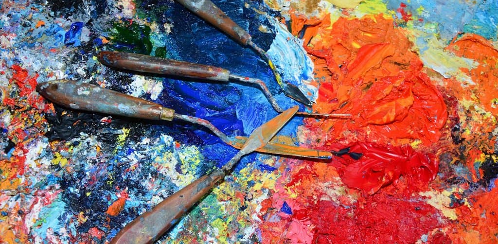 An oil painter's wooden palette photographed in close-up shows three weathered metal palette knives with cracked wooden handles laid across thick swirling impasto strokes of cobalt blue cadmium red lemon yellow and burnt umber mixed into wet sheen, the curated mixing process that mirrors how a business note broker mixes a buyer pool to find the right pricing match for each note