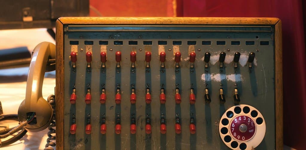 A vintage 1950s wooden-cabinet telephone switchboard with three rows of red sliding switches and a rotary dial on the right with white numerals against red ringed background sits on a paper-strewn desk beside a beige Bakelite telephone handset under warm overhead light, the analog routing equipment that symbolizes the buyer-pool routing work a business note broker does between sellers and the institutional desks that buy carry-back paper