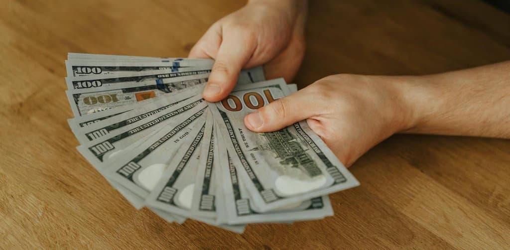 Two hands holding fanned hundred-dollar bills above a wooden desk, showing the lump-sum cash a seller receives from business note buyers