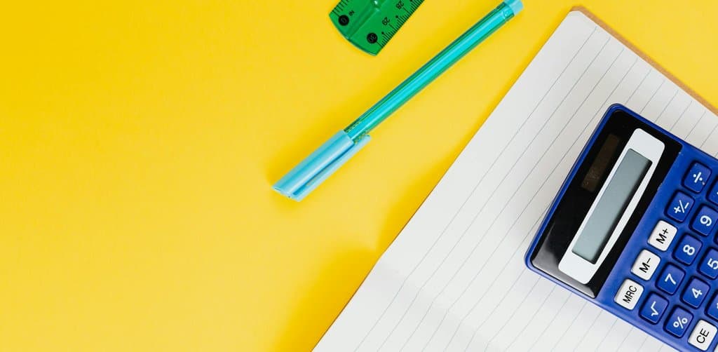 Calculator, blue pen, and lined notepad on a yellow background showing the pricing math that business note buyers run on every quote