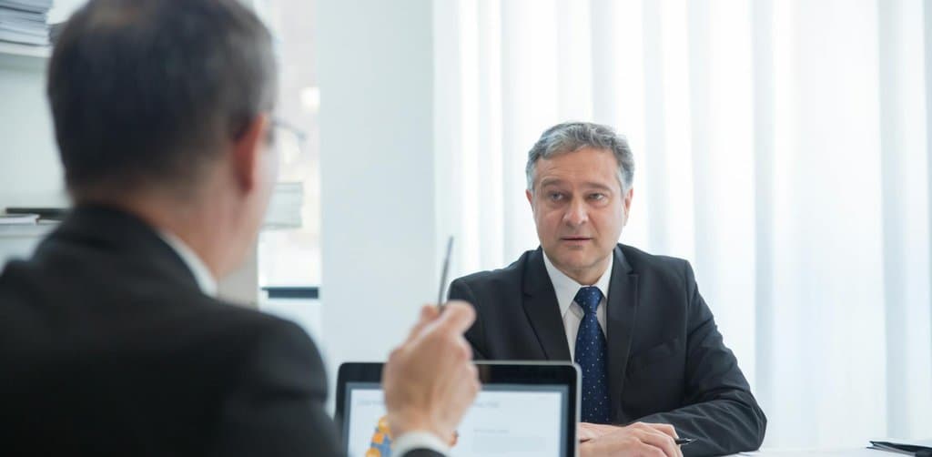 Older suited man across a desk from a younger man with laptop charts, the typical broker meeting business note buyers run with each prospective seller