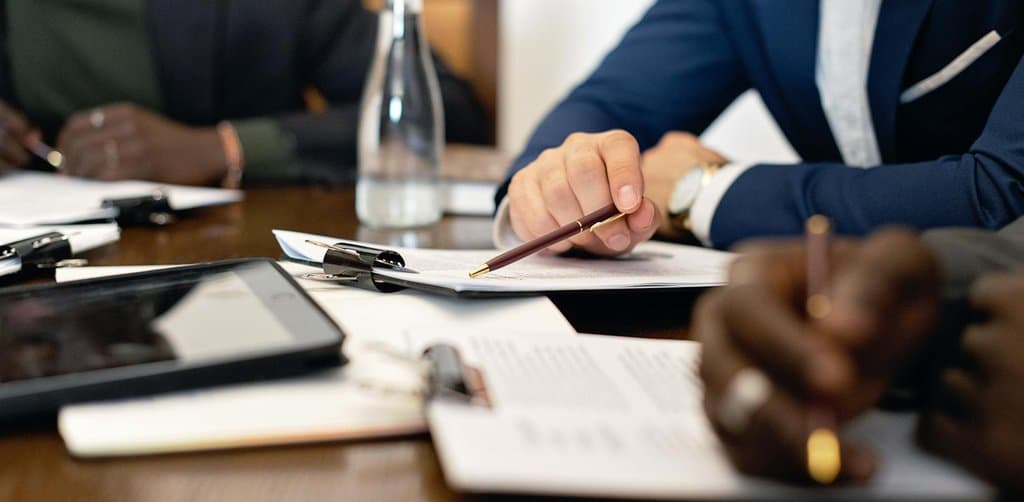 Close-up of two professionals at a wooden conference table working through documents with pens and a tablet, the buyer-seller-attorney negotiation that takes place during the diligence and LOI stage of buying a business