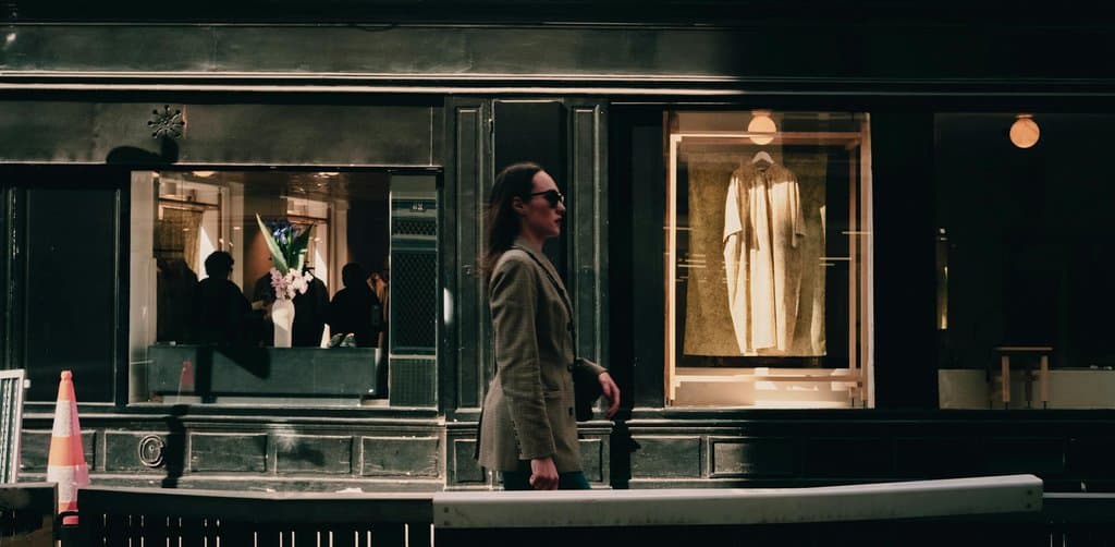 A woman in a checked blazer walking past the lit display window of a small evening boutique with garments visible behind the glass, the kind of scouting walk-through a buyer does early in the process of buying a business